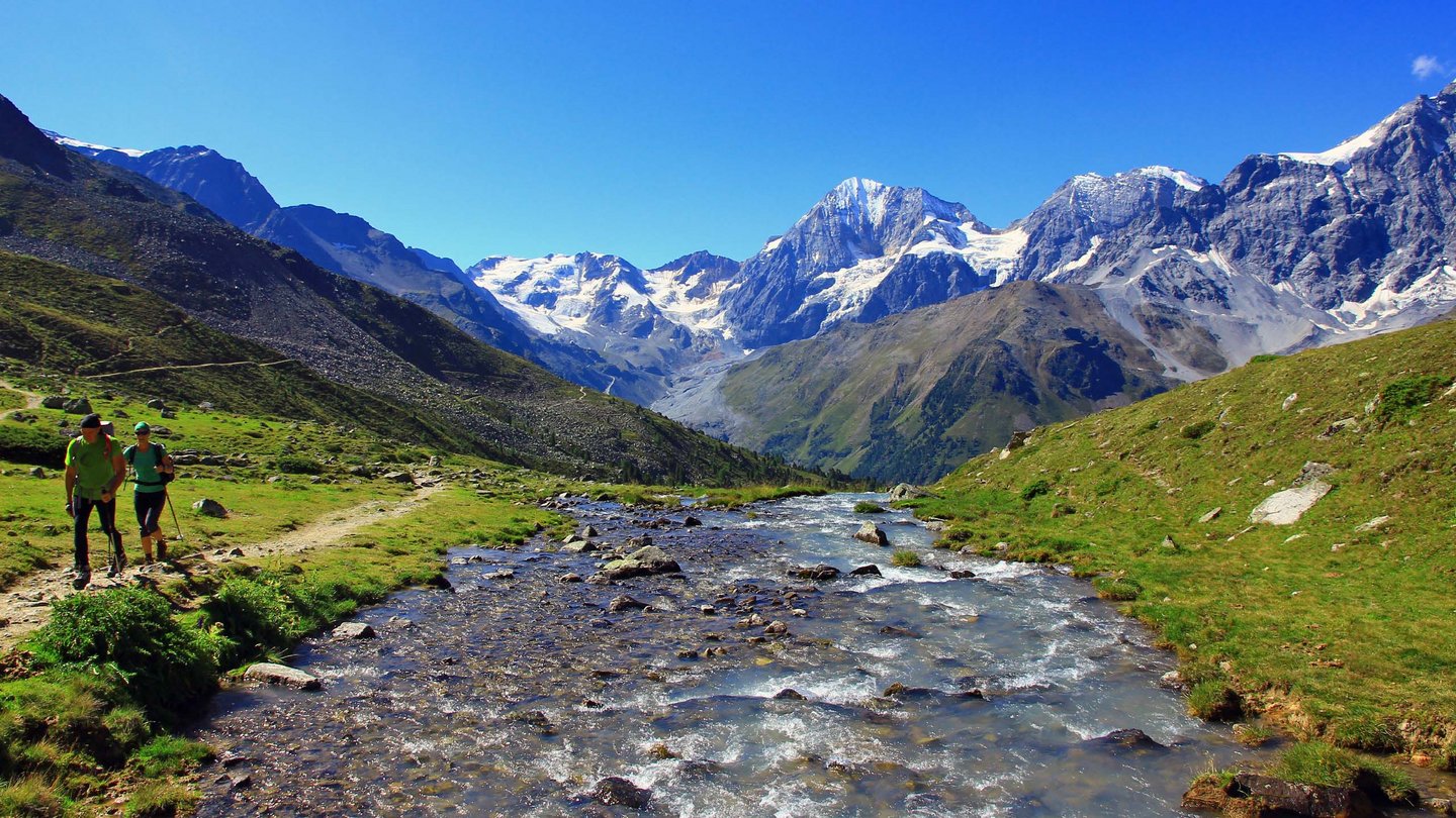 Two hikers by a mountain stream in the Stelvio National Park, surrounded by snow-capped mountains and green meadows.