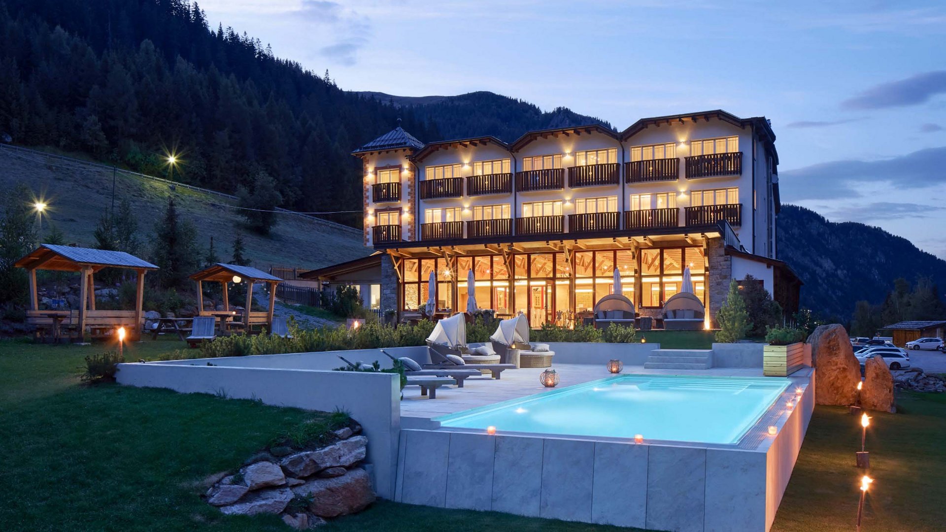 Evening view of Hotel Bella Vista with an illuminated outdoor pool in the foreground, surrounded by the picturesque South Tyrolean mountain scenery, creating a peaceful and relaxing atmosphere.