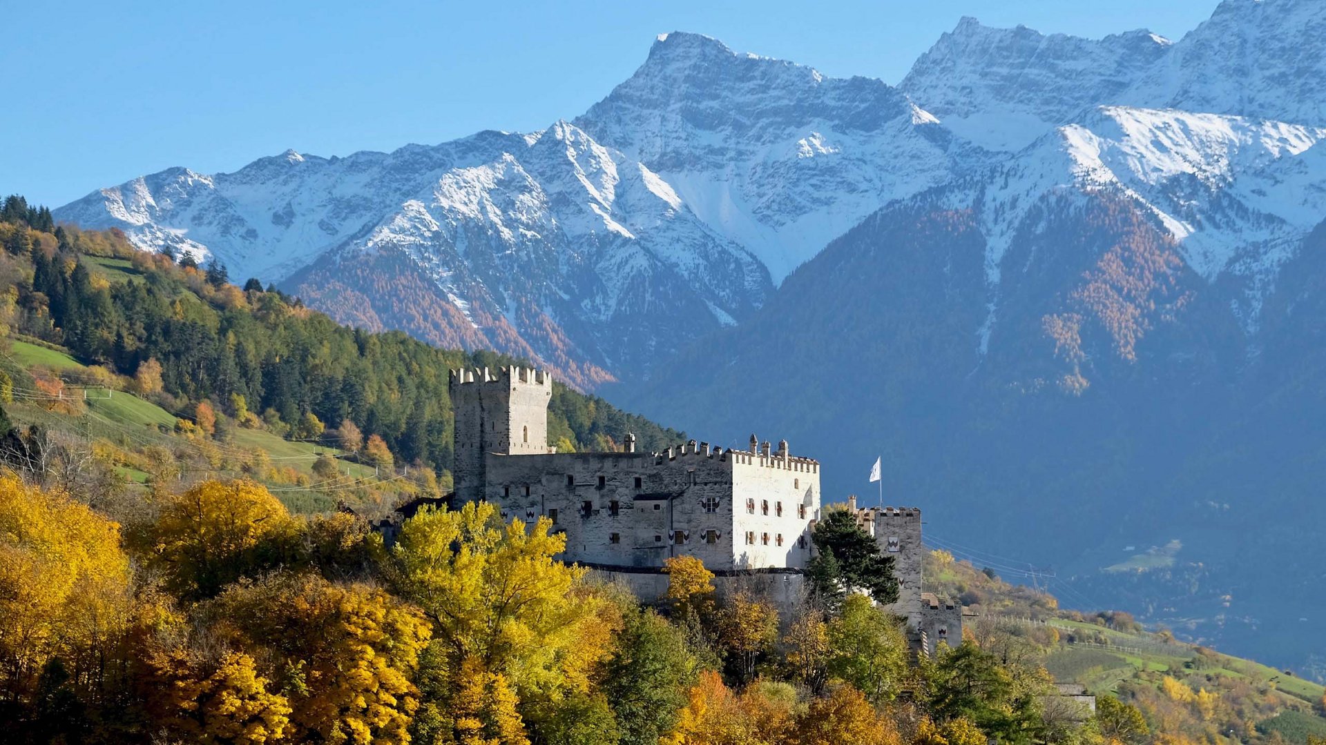 View of Churburg Castle featuring its distinctive white walls and battlements against the backdrop of the snow-capped Alps, surrounded by autumn-colored trees.