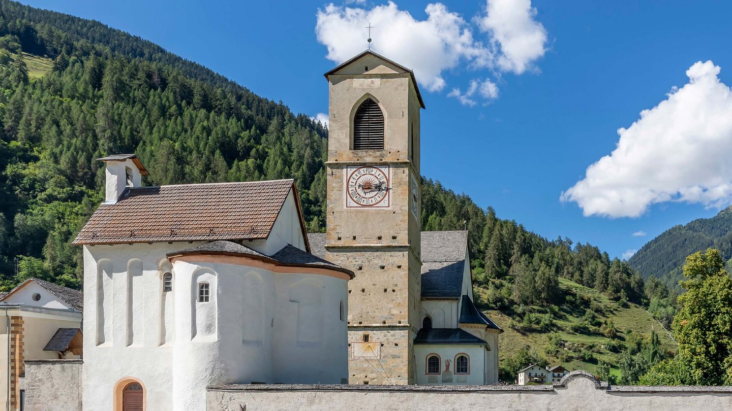 The historic St. Johann church in a sunny valley, featuring its distinctive bell tower rising against the blue sky and green mountain slopes.