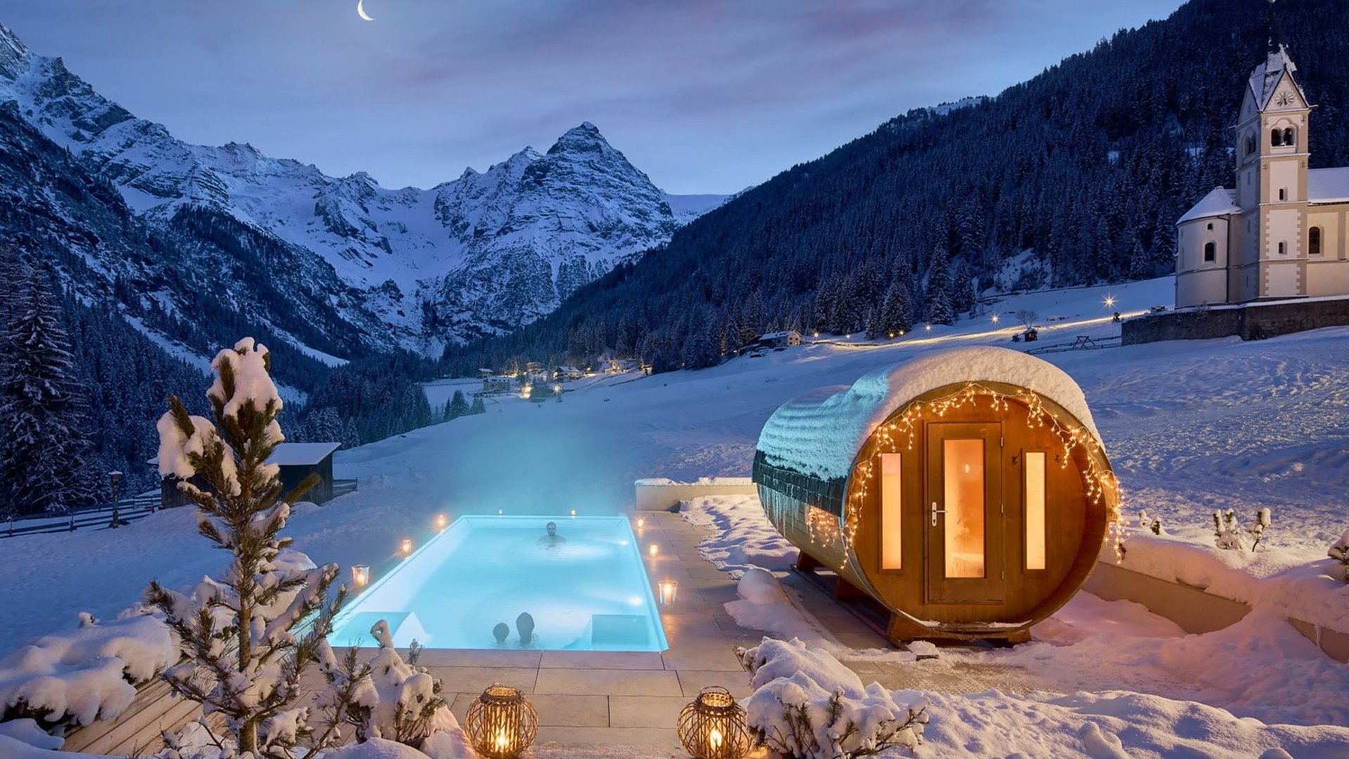 Nachtansicht des beheizten Außenpools und der angrenzenden Sauna am Hotel Bella Vista mit einer Kapelle im Hintergrund, eingebettet in die schneebedeckte Berglandschaft Südtirols.