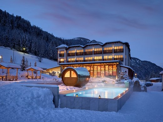 Evening view of Hotel Bella Vista in South Tyrol with lit windows and a steaming outdoor pool, surrounded by a snowy mountain landscape.